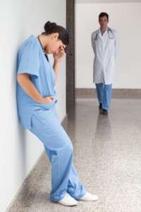 Sad nurse leans against wall in hospital corridor with doctor approaching